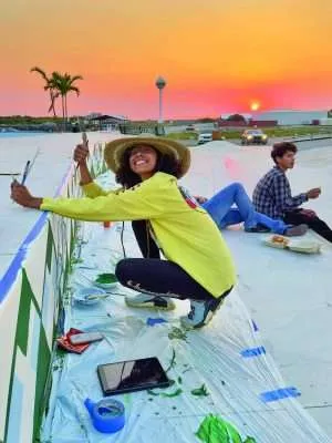 Woman painting a mural at a skate park during sunset as young people gather nearby.
