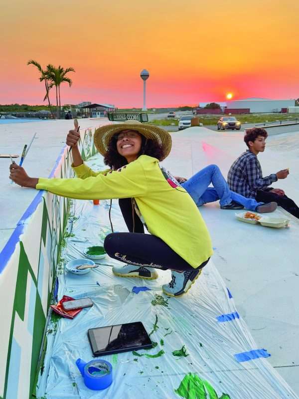 Woman painting a mural at a skate park during sunset as young people gather nearby.