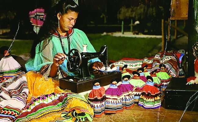 Woman sewing colorful Seminole patchwork clothing surrounded by traditional garments and dolls. Contemporary Native Americans