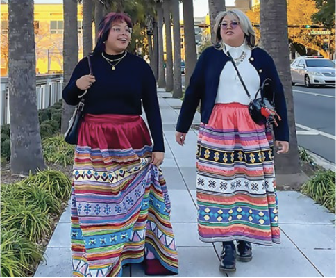 Two women wearing colorful Seminole patchwork skirts walking together on a city sidewalk. Contemporary Native Americans